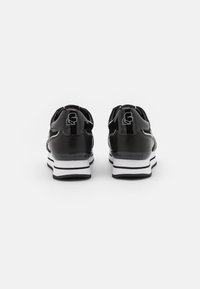 Zapatillas de charol negro con suelas blancas. Diseño redondeado que presenta un logo en el talón y rayas blancas contrastantes a lo largo de la suela.