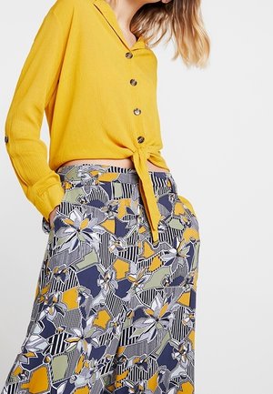 Kobieta w żółtej koszuli zapinanej na guziki, zawiązanej w pasie, oraz w spodniach z wysokim stanem o wielokolorowym kwiatowo-geometrycznym wzorze.