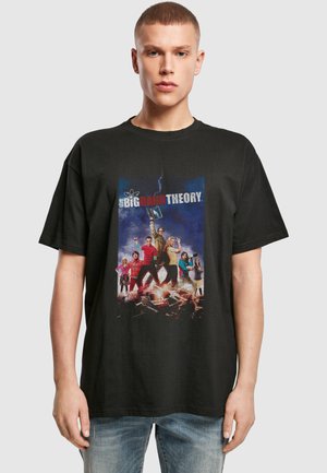 Jeune homme portant un T-shirt noir représentant le casting de The Big Bang Theory, debout sur des livres cassés avec un fond bleu orageux.