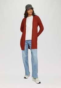Gros cardigan couleur rouille en tricot doux, porté sur un T-shirt blanc. Associé à un jean bleu clair et des baskets grises. Design simple.