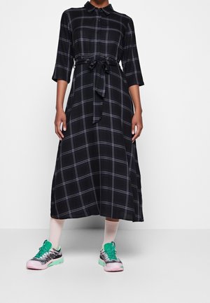 Person trägt ein schwarz-grau kariertes Midi-Hemd-Kleid mit dreiviertel Ärmeln, gebundener Taille, weißen Socken und mintgrünen Turnschuhen.