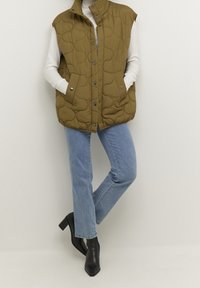 Ärmellose olivgrüne gesteppte Weste über einem weißen Langarmshirt, kombiniert mit hellblauen Jeans und schwarzen Stiefeletten mit Absatz.