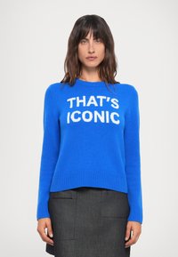 Blå strikket genser med ribbestrikkede mansjetter og kant, med stor hvit tekst "THAT'S ICONIC" på fronten, kombinert med et mørkegrått skjørt.