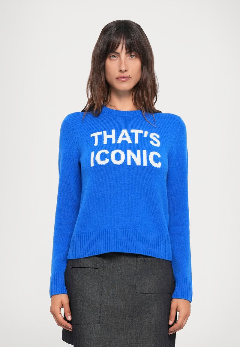 Blå strikket genser med ribbestrikkede mansjetter og kant, med stor hvit tekst "THAT'S ICONIC" på fronten, kombinert med et mørkegrått skjørt.