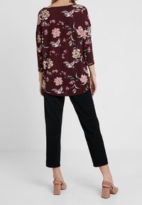 Bordeaux kleurige bloemen top met driekwart mouwen, versierd met roze bloemen en vogels. Gecombineerd met zwarte broeken en tan kleurige blokhak sandalen.