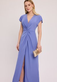 Jasnoniebieska sukienka maxi z głębokim dekoltem w kształcie litery V, krótkimi rękawami i skręconym przodem. Posiada rozcięcie do uda i złotą kopertówkę w dłoni.