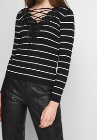 Schwarz-weißer, gestreifter Pullover mit tiefem V-Ausschnitt, Schnürung vorne und floralen Spitzenakzenten, kombiniert mit schwarzen Lederhosen.