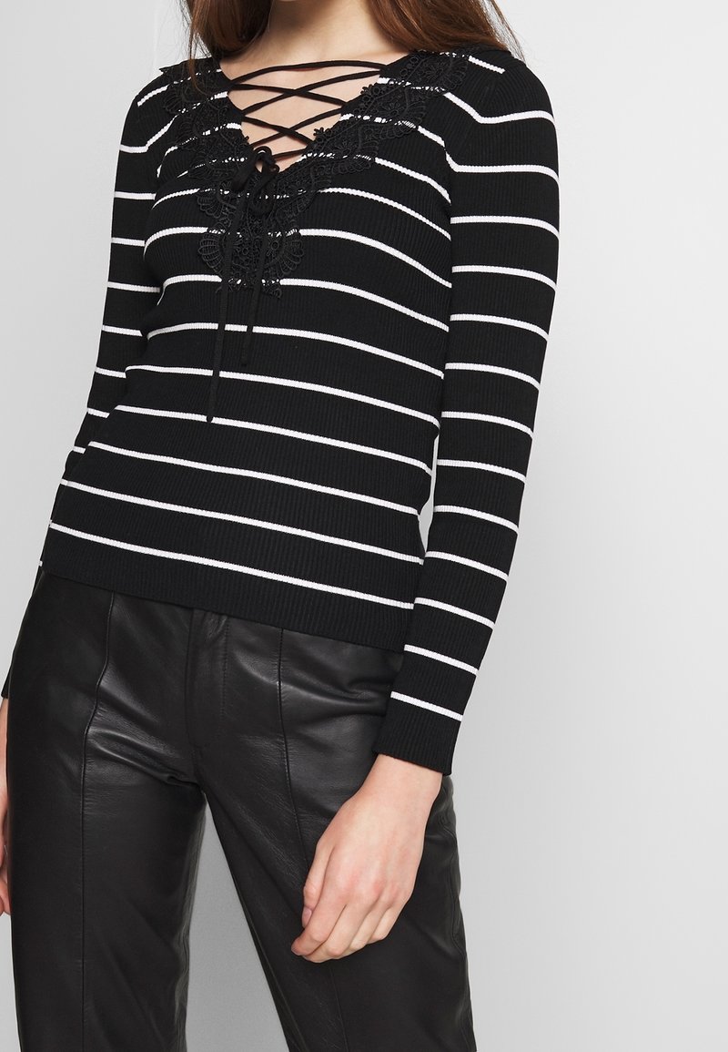 Schwarz-weißer, gestreifter Pullover mit tiefem V-Ausschnitt, Schnürung vorne und floralen Spitzenakzenten, kombiniert mit schwarzen Lederhosen.