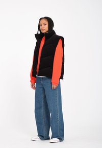 Schwarze Corduroy-Pufferweste über einem leuchtend orangefarbenen Sweatshirt, kombiniert mit lockeren blauen Jeans und weißen Sneakers. Einfaches, gepolstertes Design.