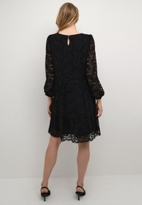 Rochie neagră din dantelă, cu mâneci lungi din material transparent, fustă evazată și detaliu cu deschidere în formă de cheie la spate. Prezintă un tiv în formă de val și un model floral texturat.