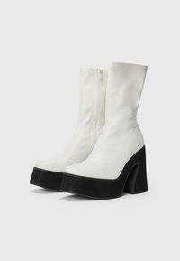 Botine albe până la gleznă, cu un finisaj din piele lucioasă, închidere cu fermoar și un toc gros negru. Textură netedă și design elegant.