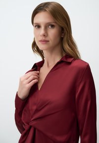 Burgundy silk blouse with a V-neck and twist knot detail at the waist. Long sleeves with buttoned cuffs. Smooth, shiny texture.