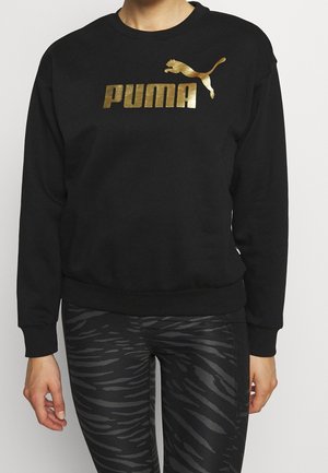 Person iført sort sweatshirt med metalisk guld Puma-logo og sorte mønstrede leggings, stående med armene afslappet ned langs siderne.