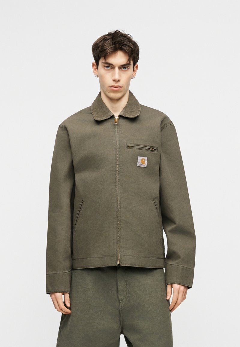 Giovane uomo con giacca verde oliva con cerniera a zip sul petto e tasca, abbinata a pantaloni, in piedi davanti a uno sfondo chiaro e semplice.