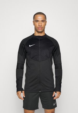 Zwarte sportjack met een rits aan de voorkant, gestructureerde mouwen en een cirkelvormig borstpaneel. Gecombineerd met bijpassende zwarte shorts met een logo.
