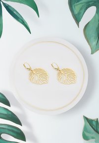 Örhängen i guldbladform med intrikata utskurna mönster hängande från lås, uppställda på en rund vit tallrik mot en ljus bakgrund.