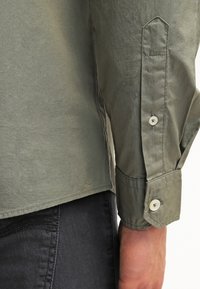 Camicia di cotone verde con maniche lunghe, polsini a bottone e colletto a punta; texture liscia e vestibilità classica, abbinata a pantaloni scuri.