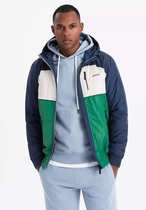 Kleurrijke jas met marineblauwe mouwen, witte en groene voorpanden, en een ritszak. Gelaagd over een lichtblauwe hoodie en bijpassende broek.