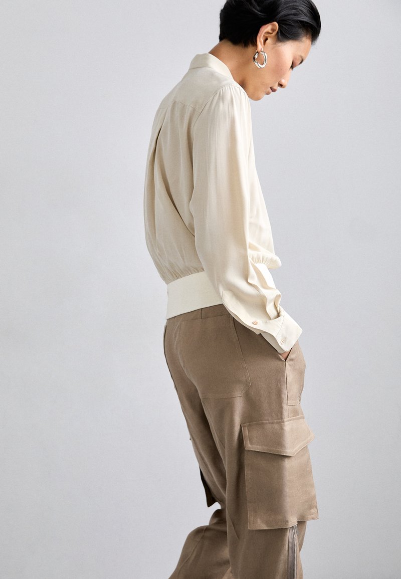 Beige Seidenbluse mit langen Ärmeln, elastischem Bund und Kragen, kombiniert mit lockeren olivfarbenen Cargo-Hosen mit Seitentaschen und sichtbaren Nähten.