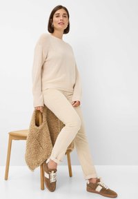 Frau in beigem Sweater und Hose sitzt auf einem Holzstuhl, hält eine braune, strukturierte Tasche und trägt braun-weiße Sneakers.