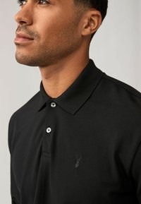 Zwarte poloshirt van een textuurstof, met drie zilveren knopen en een subtiele geborduurde logo op de linkerkant van de borst.