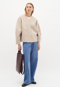 Vilnos mišinys, šviesiai pilkas švarkas su įvyniojimo dizainu, plačiomis rankovėmis ir struktūrizuota forma, derinamas su mėlynomis plačiomis kelnėmis ir ruduočiu krepšiu.