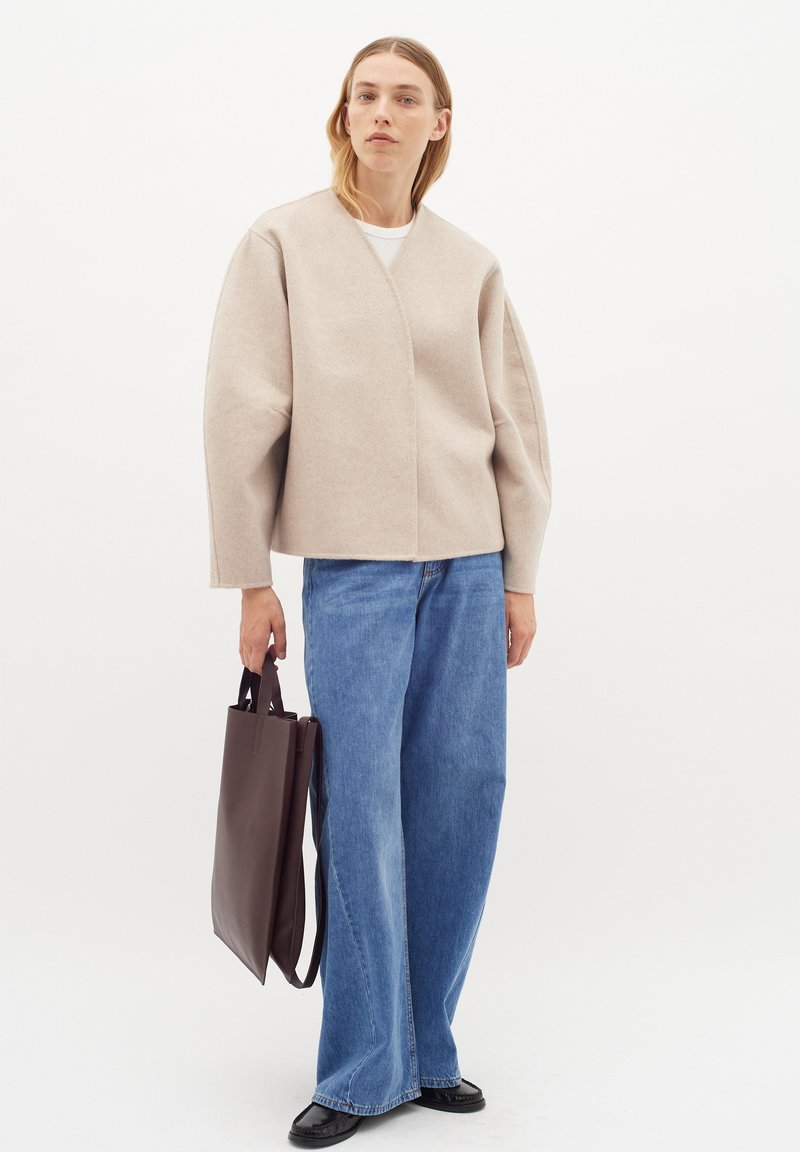 Vilnos mišinys, šviesiai pilkas švarkas su įvyniojimo dizainu, plačiomis rankovėmis ir struktūrizuota forma, derinamas su mėlynomis plačiomis kelnėmis ir ruduočiu krepšiu.