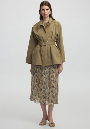 Veste ceinturée verte olive avec une coupe décontractée, associée à une robe plissée arborant un motif texturé discret dans des tons terreux. Sandales simples.