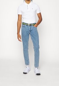Muž v bílém polo tričku s krátkým rukávem, modrých džínách s černým a žlutým páskem Tommy Jeans, bílých teniskách Nike a bílých ponožkách, stojící na bílém pozadí.