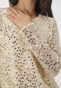 Zlatý dlouhý rukáv top s flitry, s mřížkovým vzorem, který obsahuje flitry různé velikosti v kulatém tvaru a měkkou, strukturovanou tkaninu.