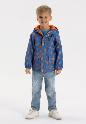 Giacca impermeabile blu con cappuccio arancione, decorata con una stampa giocosa di animali arancioni, abbinata a jeans azzurri chiari e sneaker bianche.