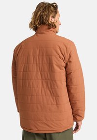 Chaqueta aislante marrón con un acabado texturizado, que presenta costuras horizontales y un cuello alto. La vista trasera muestra un diseño de ajuste relajado.