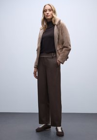 Veste en fausse fourrure marron, col roulé noir, large pantalon marron avec une poche latérale, et chaussures sombres avec des lacets blancs, sur un fond uni.