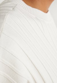 Suéter blanco de punto con textura de canalé vertical, que presenta un cuello redondo y una tela suave y ligera.