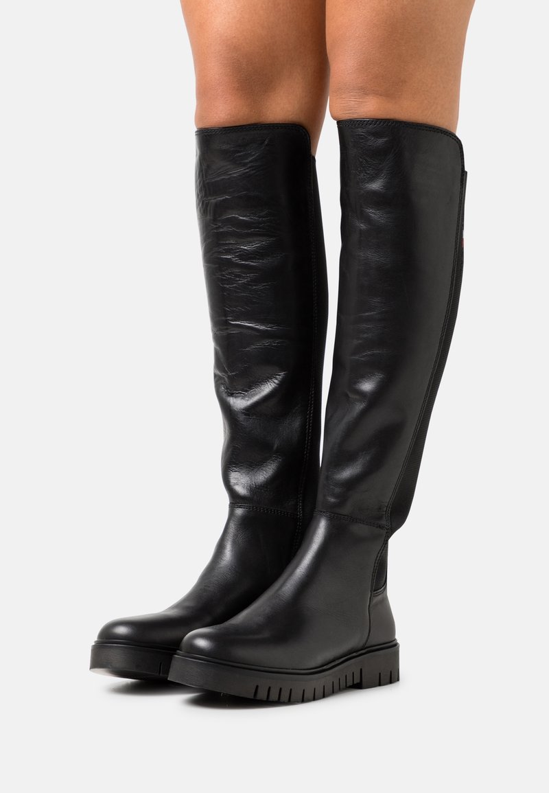 Botas de cuero negras sobre la rodilla con una textura suave, suela de goma gruesa y un ligero plataforma. Incluyen una cremallera lateral para facilitar su uso.