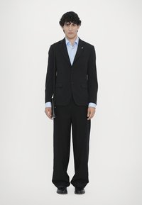 Schwarzer Anzug mit slim fit Jacke, blauer Button-up Hemd und weit geschnittenen Hosen; schwarze Lederhandtasche; Model steht vor einem weißen Hintergrund.