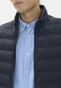 Marinblå pufferväst med quiltad textur, dragkedja och hög krage, bärs över en ljusblå skjorta med knappar i vitt.