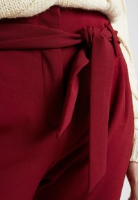 Pantalons bordeaux avec un détail de nœud noué à la taille, fabriqués dans un tissu lisse. Caractéristiques de coupe droite et accents cousus.