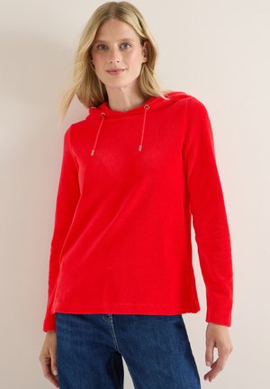 Femme blonde portant un sweat à capuche rouge vif avec des cordons et un jean bleu, se tenant devant un mur de couleur claire.