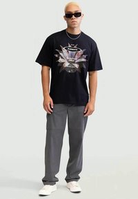 Camiseta preta com uma estampa gráfica colorida, combinada com calças cargo cinzas e ténis brancos. Os acessórios incluem óculos de sol e colares em camadas.