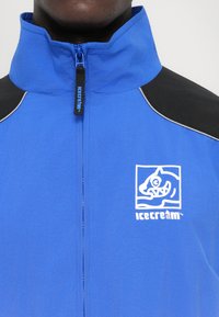 Modra zip-up jakna iz gladkega materiala. Ima črn poudarek na rami in belo logo na levi prsni strani, ki prikazuje risanega ribo.