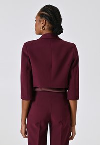 Bordeauxfarbener, taillierter Blazer mit dreiviertel Ärmeln. Glatte Stoffoberfläche mit minimalen Nähdetails. Keine sichtbaren Verschlüsse.