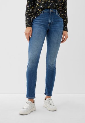 Blauwe jeans met een slim fit, voorzien van een hoge tailleband en subtiele vervaging. Gecombineerd met witte sneakers, afgebeeld tegen een effen achtergrond.