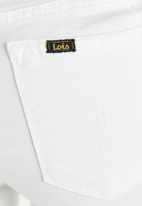 Tela de mezclilla blanca con un bolsillo trasero que presenta una etiqueta con el logotipo "Lois" en negro y dorado, mostrando una textura suave y detalles de costura clásicos.