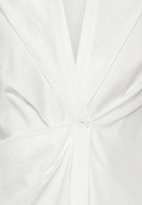 Chemise blanche avec un col en V profond, un design torsadé à l'avant et une fermeture à un bouton transparent à la taille. Texture de tissu lisse.