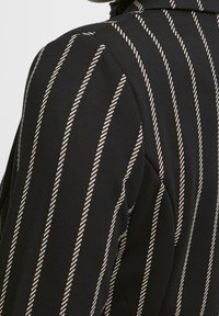 Tissu noir avec des rayures verticales beiges rappelant une corde, présentant une texture lisse et une coupe ajustée, mettant en valeur une silhouette structurée.