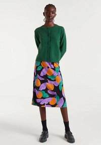 Mujer de pie con un cárdigan verde con botones, mocasines negros y una falda colorida con patrones de hojas en naranja, púrpura y verde.