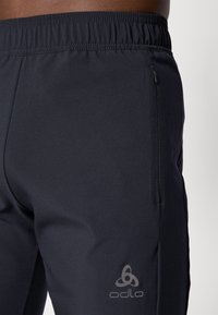Sorte treningsbukser laget av glatt, elastisk stoff med strikket liv. Har en glidelåslomme og Odlo-logo på låret.