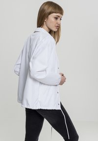 Jachetă ușoară albă, cu închidere cu nasturi, talie elastică și mâneci lungi. Textură netedă, fără modele sau accente vizibile.