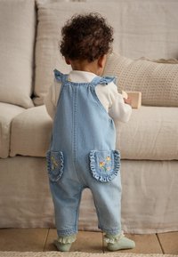 Salopette en denim bleu clair, avec des bretelles froncées, des accents de fleurs brodées sur les poches et des chaussettes vertes texturées avec un bord en dentelle.